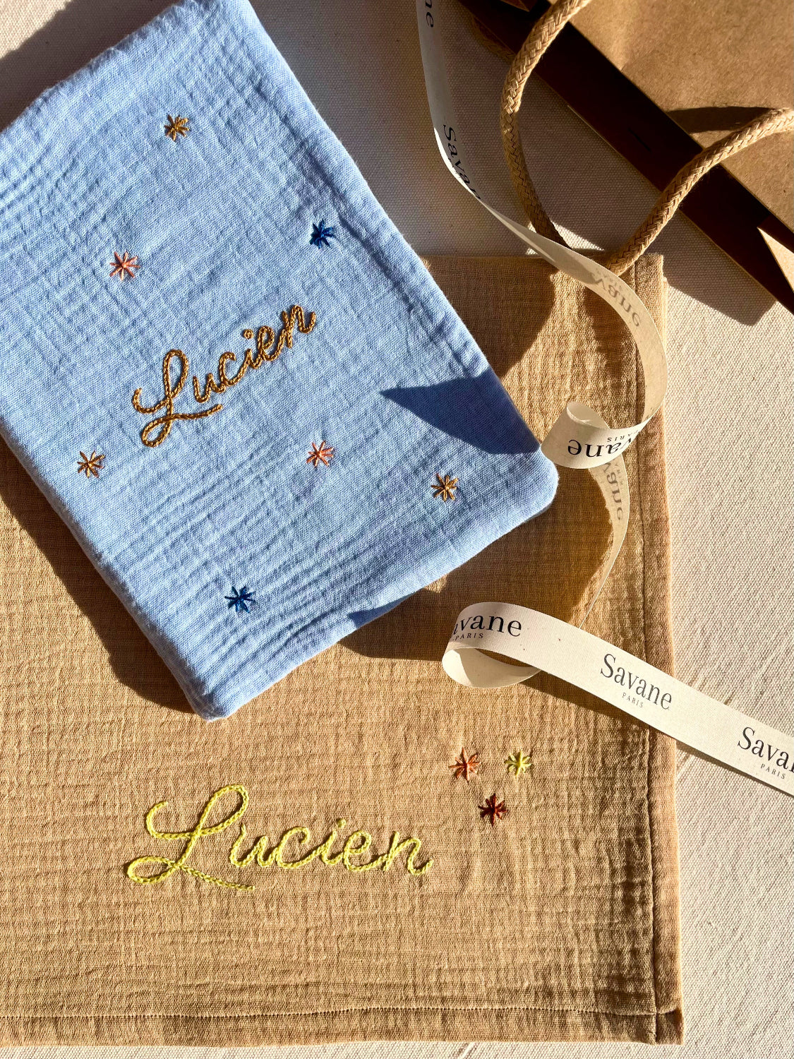 Cadeau de naissance composé d’un lange beige brodé et d’un protège carnet de santé, présenté dans un emballage cadeau premium Savane Paris.