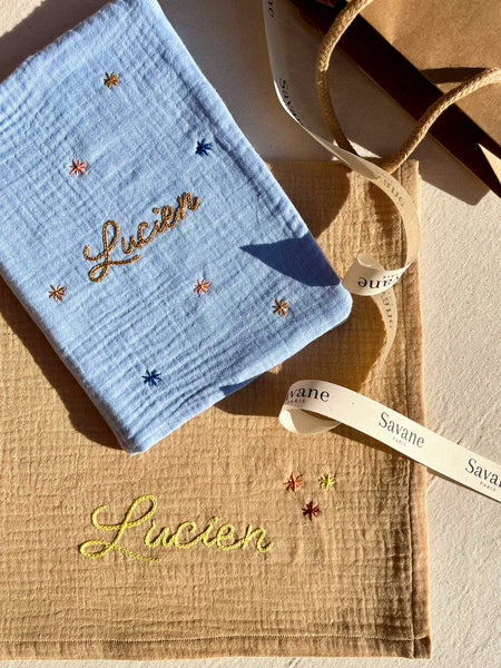 Cadeau de naissance composé d’un lange beige brodé et d’un protège carnet de santé, présenté dans un emballage cadeau premium Savane Paris.