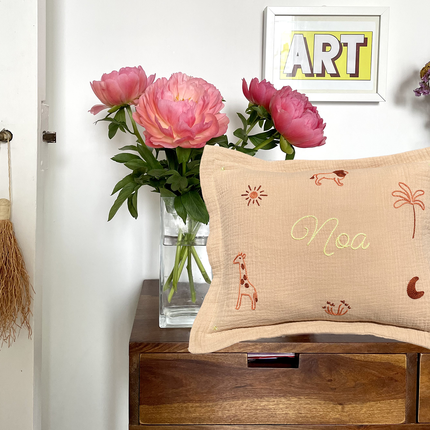 Coussin de naissance beige brodé main avec le prénom Noa et motifs savane, posé sur un meuble en bois avec bouquet de pivoines – création artisanale Savane Paris.