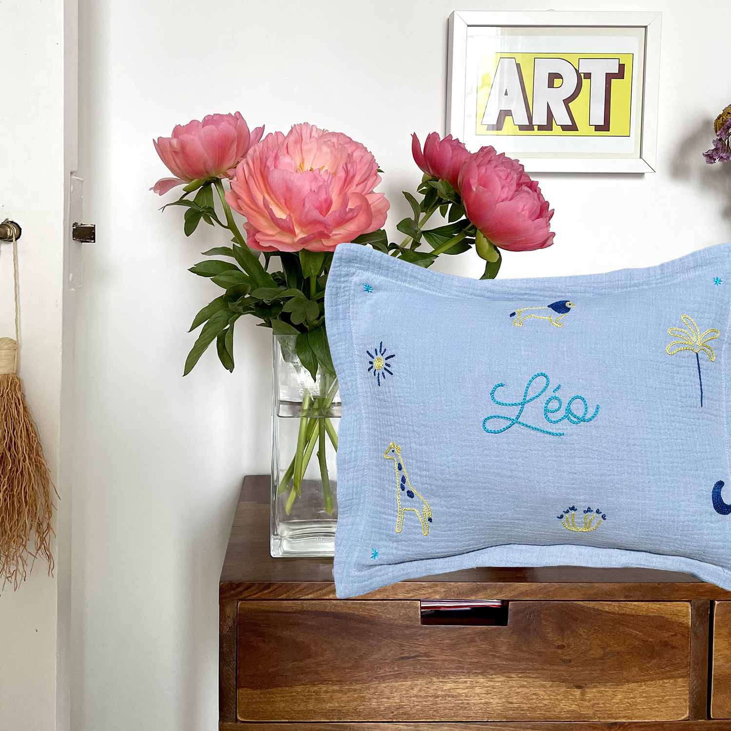 Coussin de naissance bleu brodé main avec le prénom Léo et motifs savane, présenté sur une console en bois avec bouquet de pivoines – création faite en France par Savane Paris.