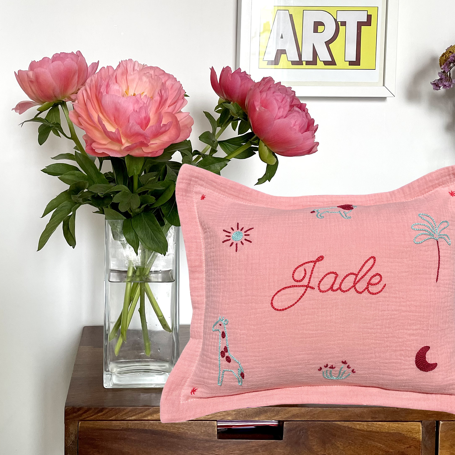 Coussin de naissance rose brodé main au prénom Jade avec motifs girafe, soleil et palmier, photographié sur un meuble en bois avec fleurs – création artisanale française Savane Paris.