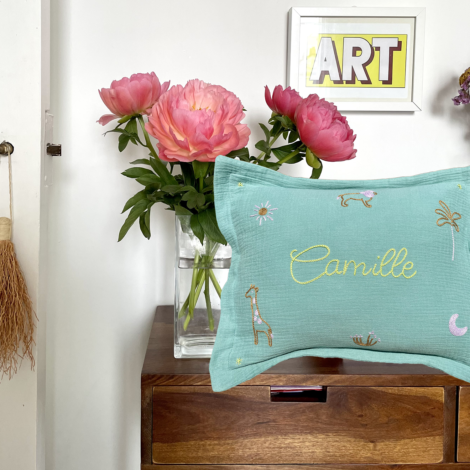 Coussin de naissance vert brodé main au prénom Camille avec motifs girafe, soleil et palmier, mis en scène sur un meuble avec fleurs – création artisanale française Savane Paris.