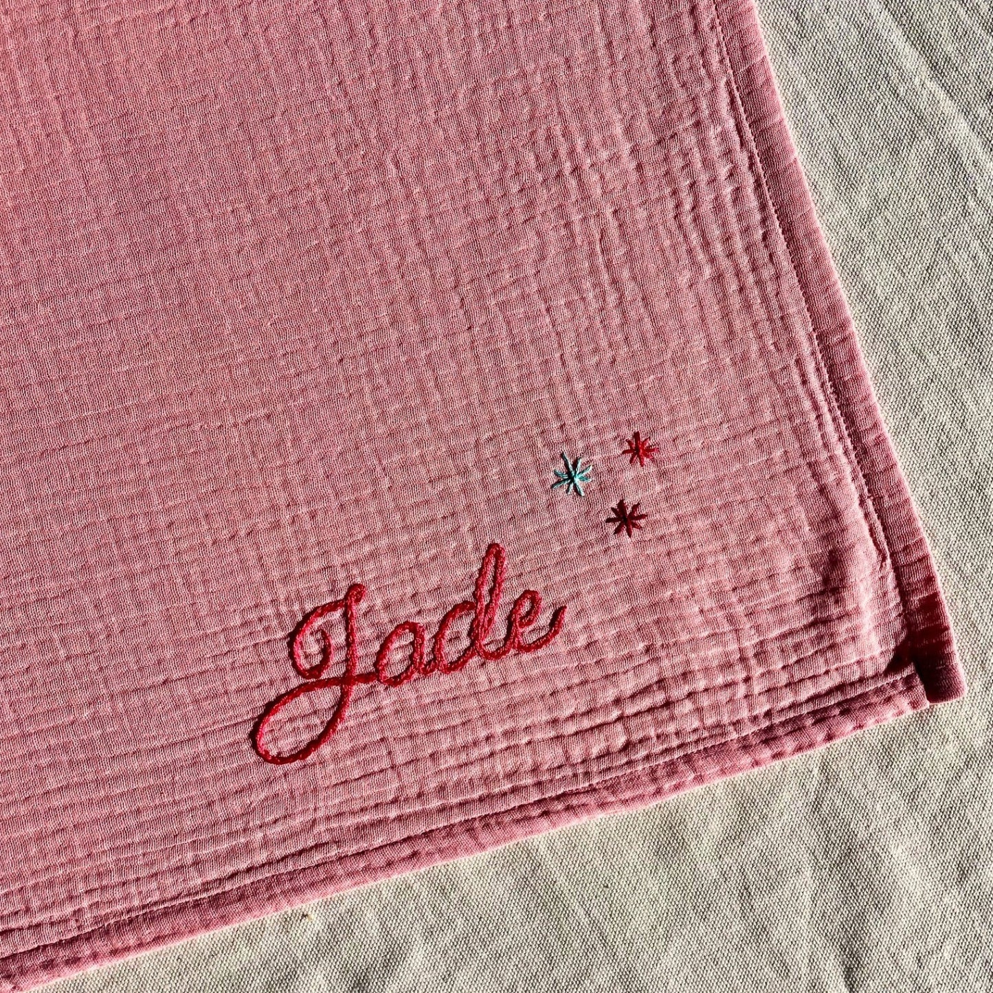 Détail de la broderie d’un lange bébé rose en gaze de coton, personnalisé au prénom et orné de petites étoiles, brodé à la main en France par Savane Paris.