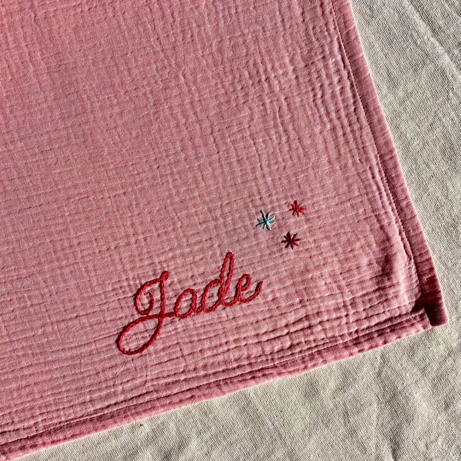 Détail de la broderie d’un lange bébé rose en gaze de coton, personnalisé au prénom et orné de petites étoiles, brodé à la main en France par Savane Paris.