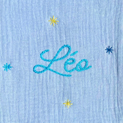 Zoom sur la broderie du prénom réalisée à la main sur un protège carnet de santé bleu cielSavane Paris – fil DMC, savoir-faire artisanal français, cadeau de naissance personnalisé.